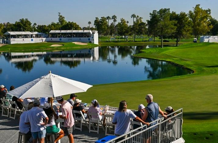 Spectators gathered on an outdoor patio overlooking a golf course pond, green fairways, and white tournament hospitality tents beyond.