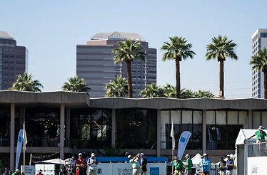 Golfer teeing off at a PGA Tour Champions event, with Charles Schwab banners, spectators, palm trees, and a city skyline behind.
