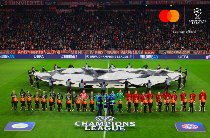 Players lined up for UEFA Champions League anthem ceremony, giant star-ball banner displayed across pitch before packed stadium.