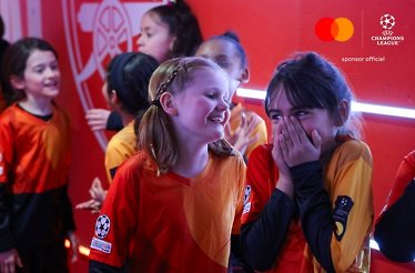 A group of young girls, wearing UEFA orange and red kit, stand together and smiling, at the walkout tunnel. 
