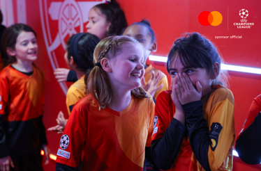 Young UEFA Champions League mascots in orange kits smiling and posing together in the player tunnel pre-match.