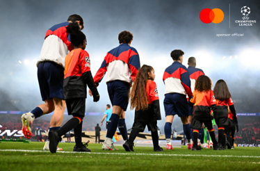 Players and child mascots walk out from the tunnel toward the pitch at a UEFA Champions League stadium, image captured from the below ground.