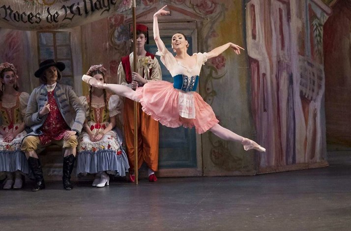 Dancer leaps gracefully in village festival setting with costumed onlookers and painted backdrop.