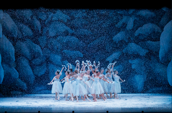 A group of ballet dancers in white tutus pose with raised arms as artificial snow falls on a blue, ice-like stage set.
