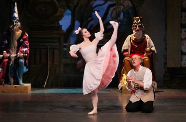 Ballet dancer in pink costume performs on stage with theatrical set and seated characters.