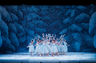 A group of ballet dancers in white tutus pose with raised arms as artificial snow falls on a blue, ice-like stage set.