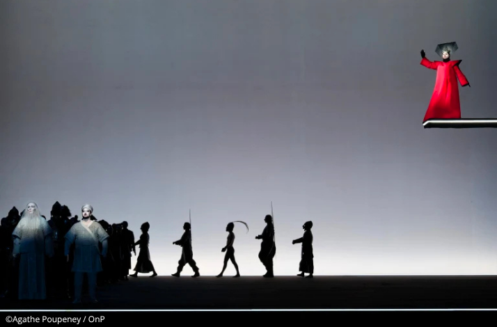 Silhouetted cast moving across the stage below Princess Turandot in red at the Opéra Bastille.