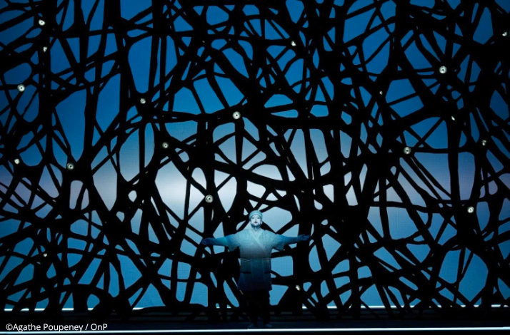 An opera singer performing with arms outstretched behind an intricate black lattice screen at the Opéra Bastille.