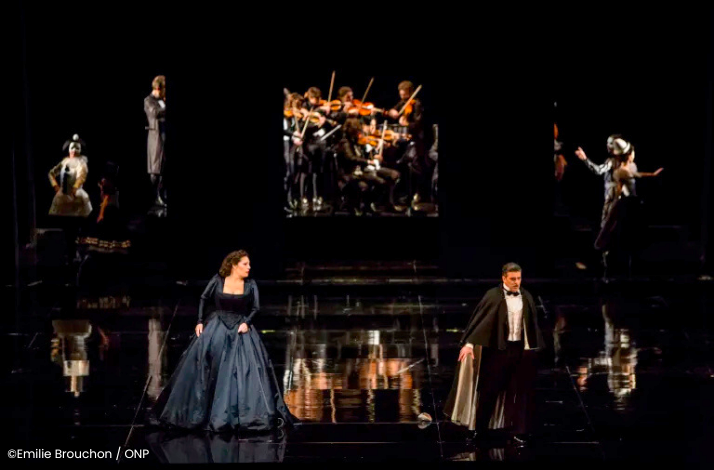 Opera stage with two performers in formal costumes and an orchestra projected in the background. ©Emilie Brouchon / ONP