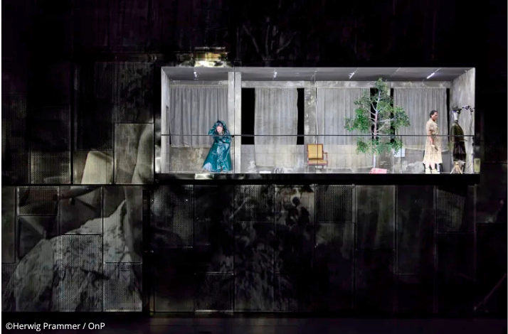 Two performers on an elevated stage set with sparse furnishings and a bare tree, during Wagner's The Valkyrie at Opera Bastille.. © Herwig Prammer / OnP.