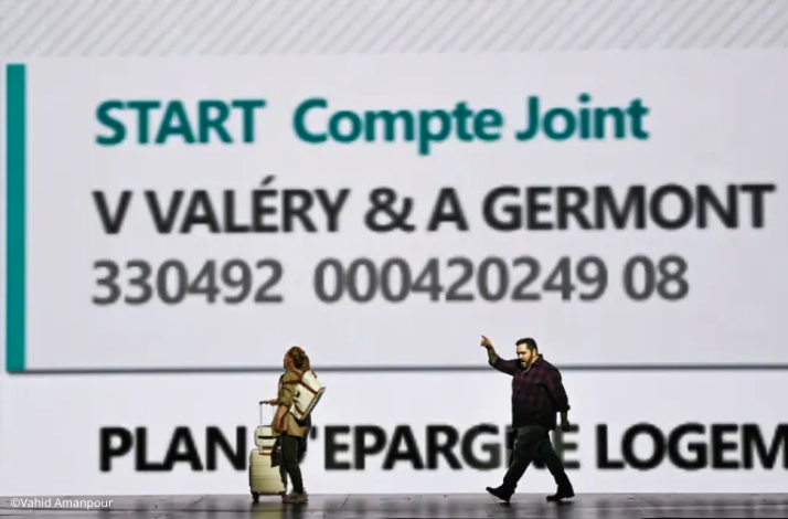 Two performers walk in front of a giant projected account screen showing the names V. Valéry and A. Germont. © Vahid Amanpour