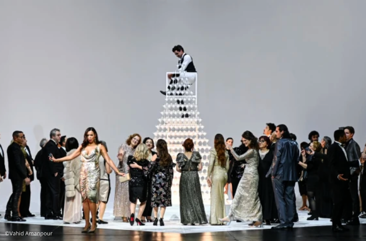 A group of elegantly dressed people gather around a tall pyramid of glasses, with a person standing on top. © Vahid Amanpour