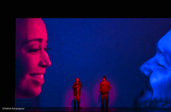 Two performers stand on stage under dramatic red and blue lighting, with large projected faces in the background.© Vahid Amanpour