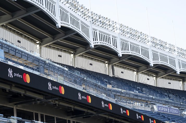 A view of a baseball stadium’s upper deck with empty seats and bright light rigs, featuring a ribbon board repeating NY prefers and an ASPEN VODKA sign.