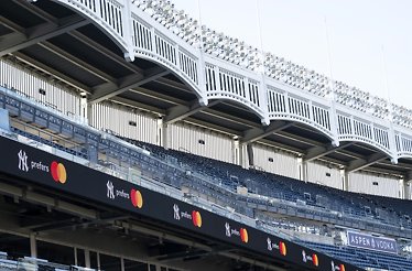 A view of a baseball stadium’s upper deck with empty seats and bright light rigs, featuring a ribbon board repeating NY prefers and an ASPEN VODKA sign.