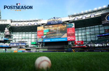 Baseball resting on American Family Field grass, with the stadium scoreboard and MLB Postseason 2026 logo visible in upper-left corner.
