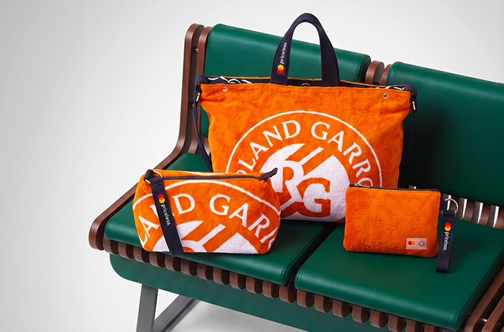 Three orange Roland-Garros upcycled towel accessories displayed on green stadium seating: large tote bag, medium crossbody bag, and small pouch, all featuring the RG logo in white.
