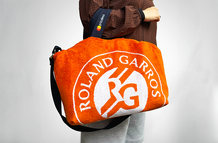 Person carrying orange Roland-Garros crossbody bag made from upcycled tournament towels, featuring the iconic RG logo in white terry cloth with black adjustable strap.