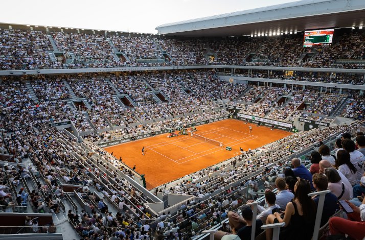 A fan's wide view of a live tennis game in Roland-Garros stadium, filled with fans and an open telescopic roof.