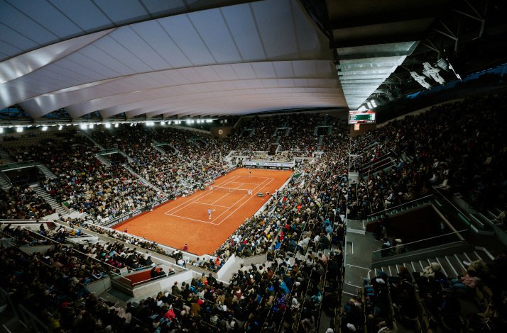 A fan's wide view of a live tennis game in Roland-Garros stadium, filled with fans and an open telescopic roof.