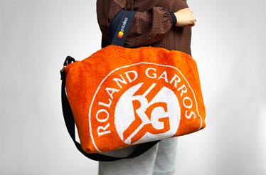 Person carrying orange Roland-Garros crossbody bag made from upcycled tournament towels, featuring the iconic RG logo in white terry cloth with black adjustable strap.
