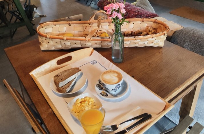 Breakfast tray with a cappuccino, sandwich, scrambled eggs, and orange juice on a wooden café table.