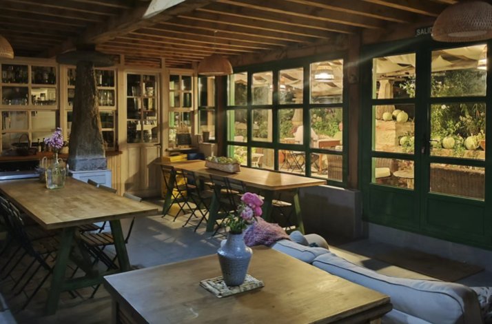 Cozy rustic interior with exposed wooden beams, long wooden tables, pendant lights, and large green-framed windows looking out onto a garden patio.