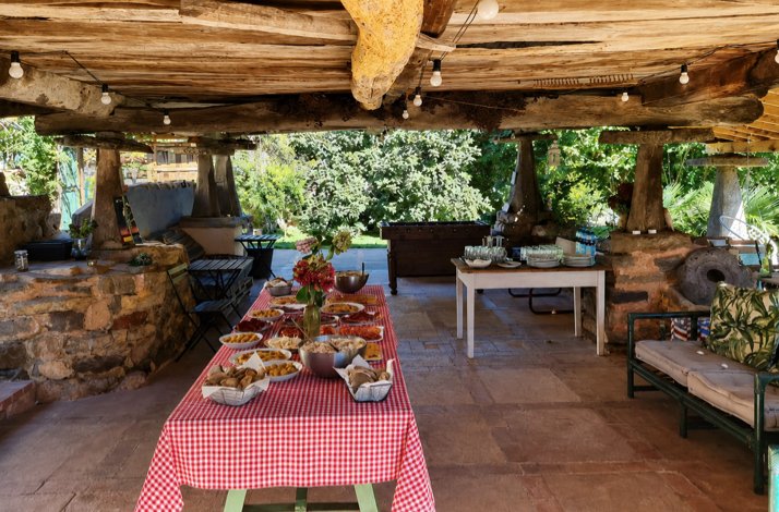A rustic open-air patio with stone walls and a wooden-beam ceiling, featuring a long table covered with a red gingham cloth and bowls of food.