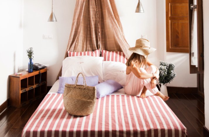 Woman relaxing on a striped bed with a straw hat and bag in a cozy, airy bedroom.