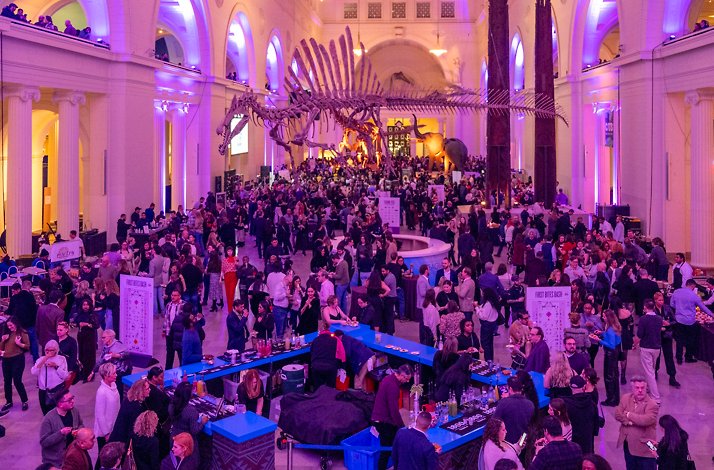 Large crowd enjoying food and drinks at a vibrant event under a dinosaur skeleton in a grand hall.