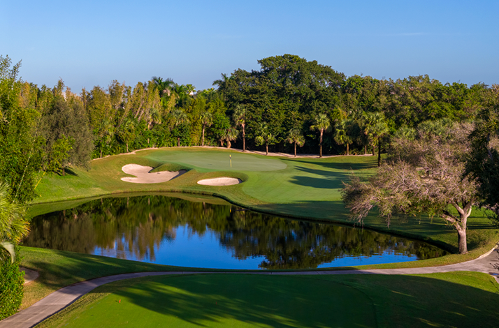 A sunlit golf course green with a flagstick, sand bunkers, and a reflecting water hazard.