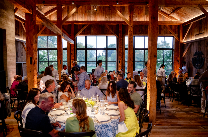 Guests dining at round tables inside a rustic wooden barn venue with large windows.