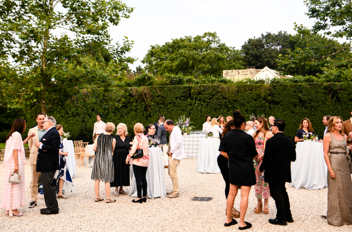 Guests mingling at an outdoor cocktail party on a gravel patio with green hedges.