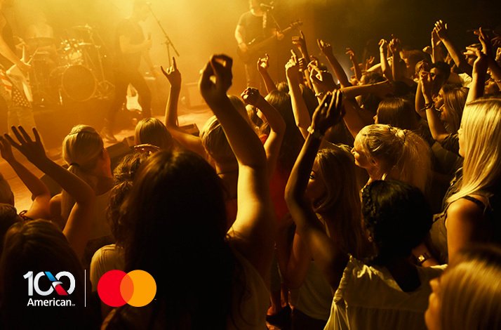 Concert crowd with hands raised in a music stadium. Logos of American Airlines and Mastercard in the bottom left corner.
