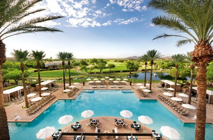 Usage statistics Luxurious resort pool area surrounded by palm trees, sun loungers, and white umbrellas, with views of green golf courses and a pond under a bright blue sky.