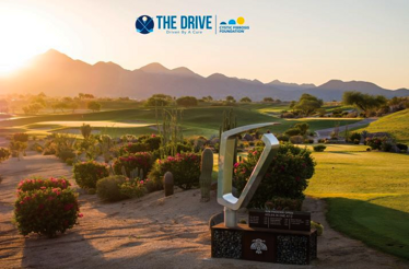 A scenic golf course at sunrise with desert landscaping, mountains in the background, and a modern trophy sculpture in the foreground, featuring The Drive and Cystic Fibrosis Foundation logos.