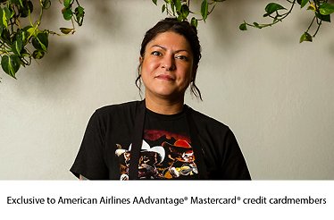 Chef Misti Norris wearing a black graphic tee and apron, posed in front of a neutral wall with trailing greenery overhead.