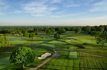 Aerial view of a lush golf course with sand bunkers, fairways, and trees under a blue sky.