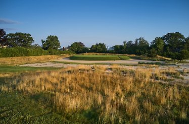 Golf course with a raised green, surrounded by sand bunkers and tall grass under a clear blue sky.
