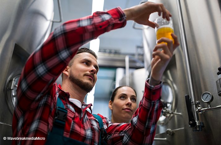 Two brewers inspecting a glass of beer in front of large brewery tanks at Spatenbrauerei Oktoberfest. @WavebreakmediaMicro
