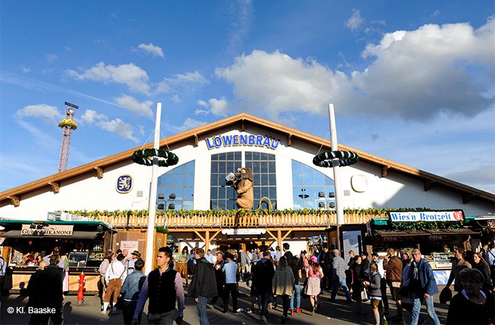 Festivalgoers walking outside the Löwenbräu beer tent at Oktoberfest, featuring the 'Löwenbräu' sign and a lion statue above the entrance. © Kl. Baaske