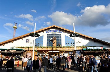 Festivalgoers walking outside the Löwenbräu beer tent at Oktoberfest, featuring the 'Löwenbräu' sign and a lion statue above the entrance. © Kl. Baaske