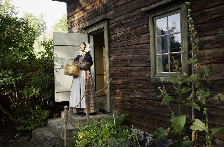 A mannequin in a Swedish traditional dress standing by an open door of one of the buildings of the Skansen museum