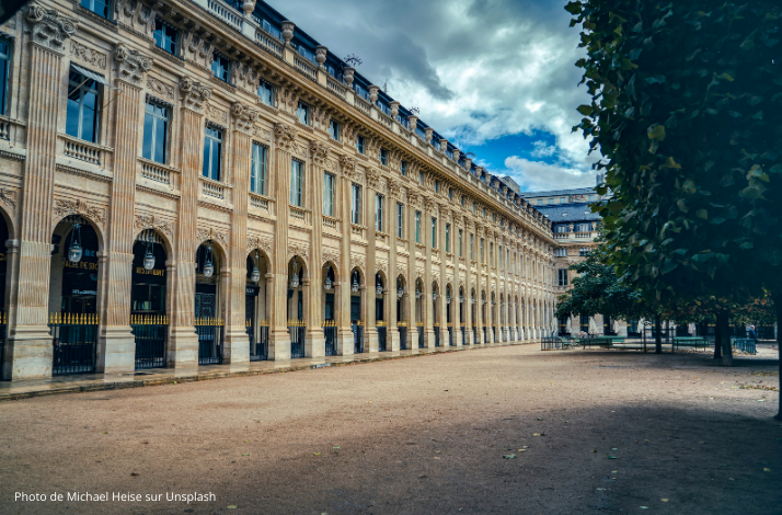 Elegant façade of Palais Royal with arched windows and a tree-lined courtyard under a dramatic sky.