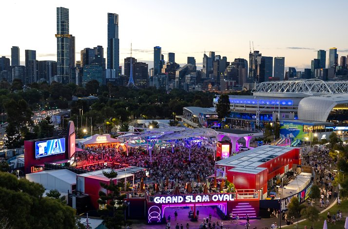 Dusk view of Melbourne skyline behind a brightly lit Australian Open precinct with crowds and the “Grand Slam Oval” entrance.