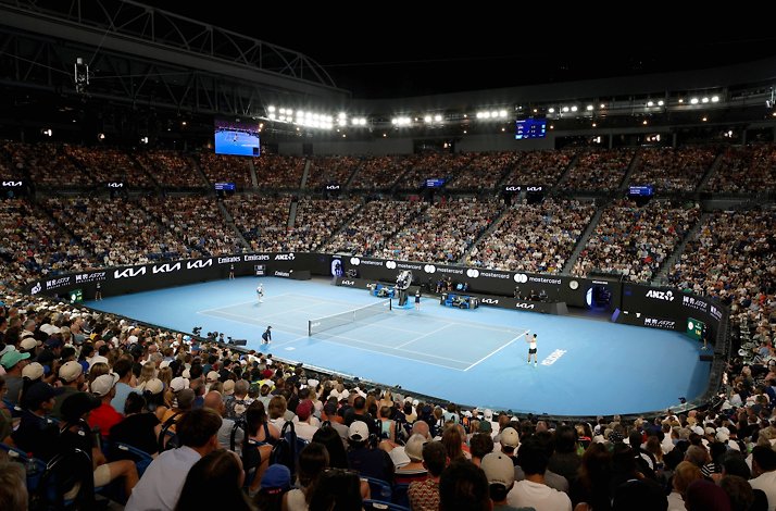 Wide interior view of a packed tennis arena during a match on a blue hard court under bright stadium lights.
