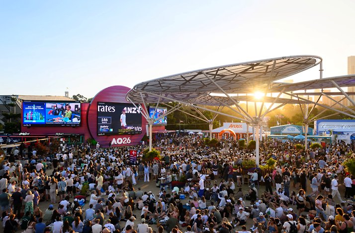 Large crowd gathers at the Australian Open fan zone at sunset, with big screens and canopy structures overhead.