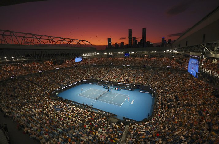 Night match in a sold-out tennis stadium with a blue court, city skyline silhouetted against a red-orange sunset.