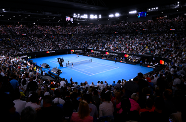 A packed tennis arena during a match at the Australian Open, with the blue court illuminated and a vast audience.