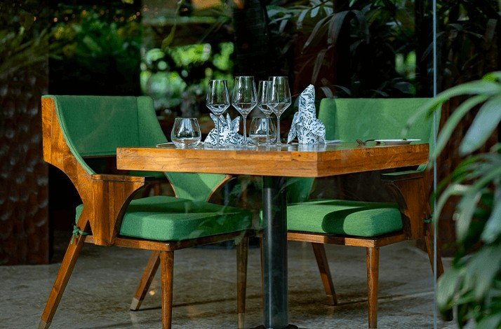 A wooden table prepared with tableware sets along with green chairs in a garden patio setting at Mozaic Bali restaurant.